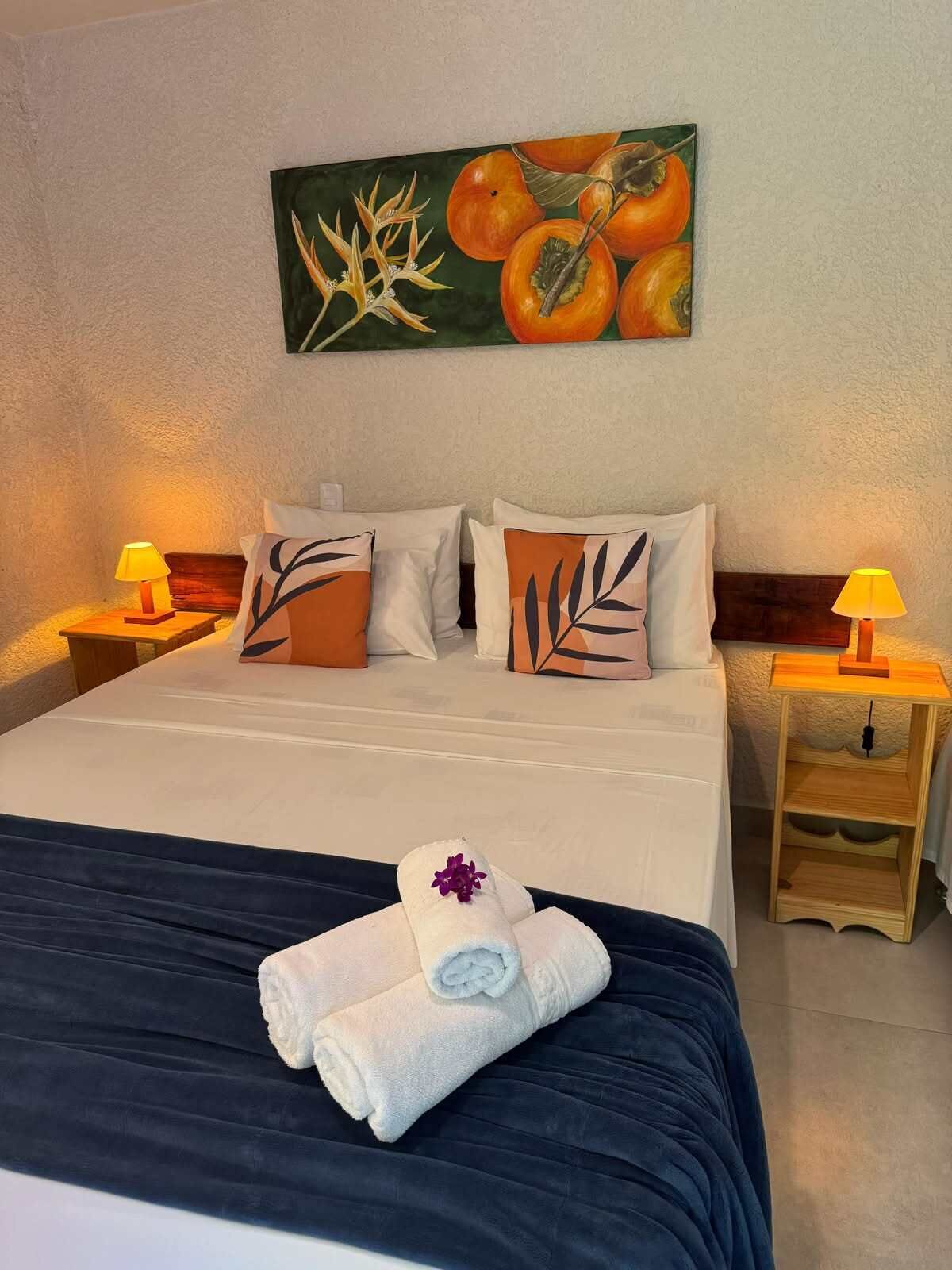 Uma cama bem arrumada com um cobre-leito azul escuro, duas almofadas com estampas de folhas, um quadro na parede com tema de frutas e duas luminárias de cabeceira.