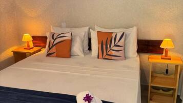 Uma cama bem arrumada com um cobre-leito azul escuro, duas almofadas com estampas de folhas, um quadro na parede com tema de frutas e duas luminárias de cabeceira.