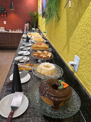 A buffet with various desserts and a long table with plates and utensils. - Pousada Preciosa do Arraial - com Natura e L’Occitane - recém inaugurada (Porto Seguro)