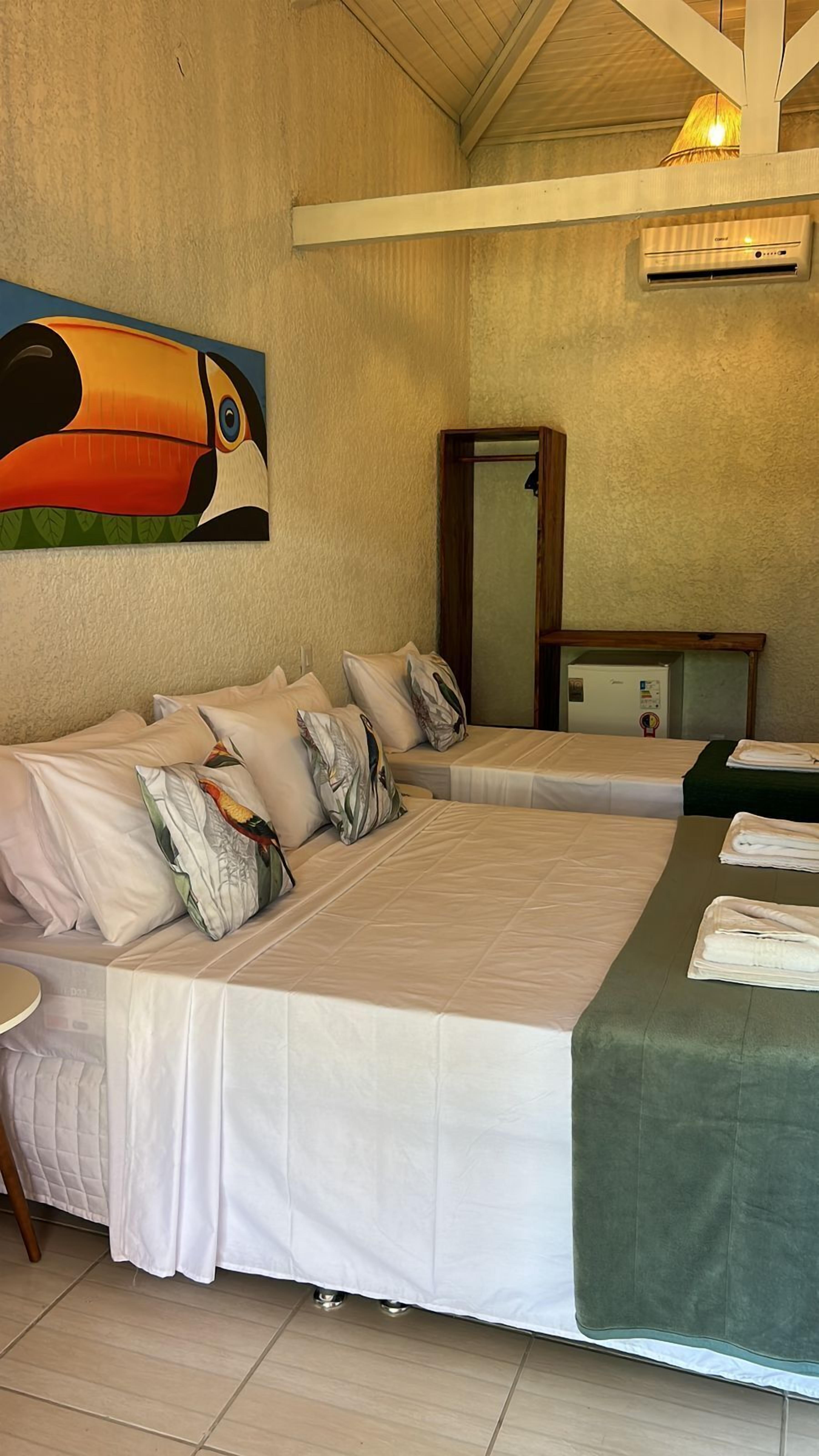 Une chambre d’hôtel avec un grand lit, un plus petit lit, un tableau représentant un toucan, un miroir et un ventilateur de plafond.