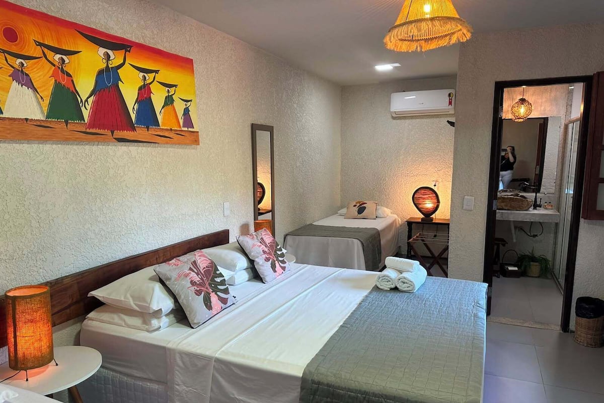 Habitación de hotel con cama, mesitas de noche, un cuadro, aire acondicionado y baño visible a través de una puerta abierta.
