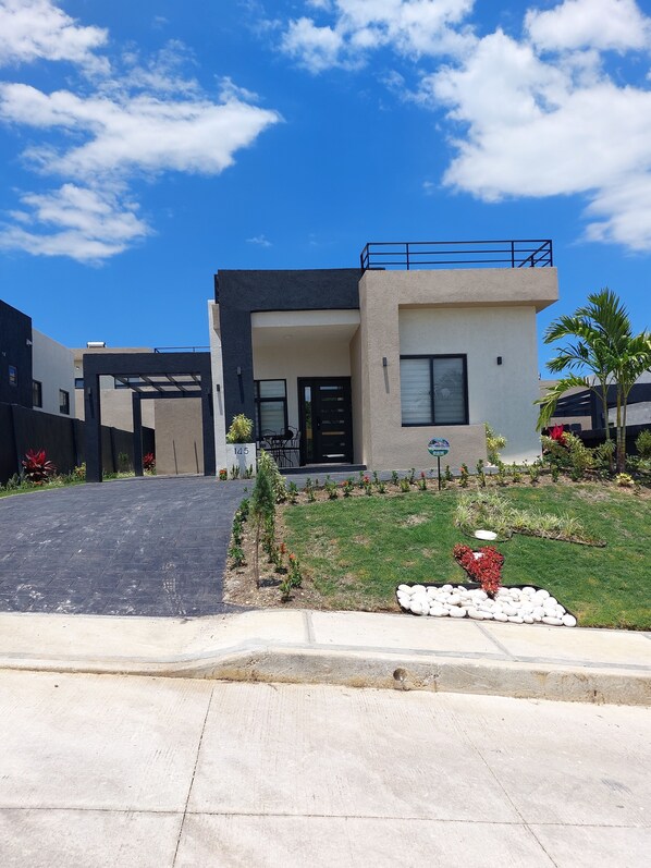 A modern two-story house with a flat roof, a balcony, and a well-maintained garden with various plants and a small pond.