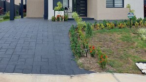 Une maison moderne à deux étages, dotée d’une allée pavée, d’un portail noir et d’un petit jardin agrémenté de fleurs rouges.
