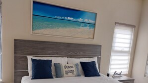 Une chambre à coucher avec un grand lit, une tête de lit, un tableau représentant une plage, une fenêtre avec des stores et un ventilateur de plafond.