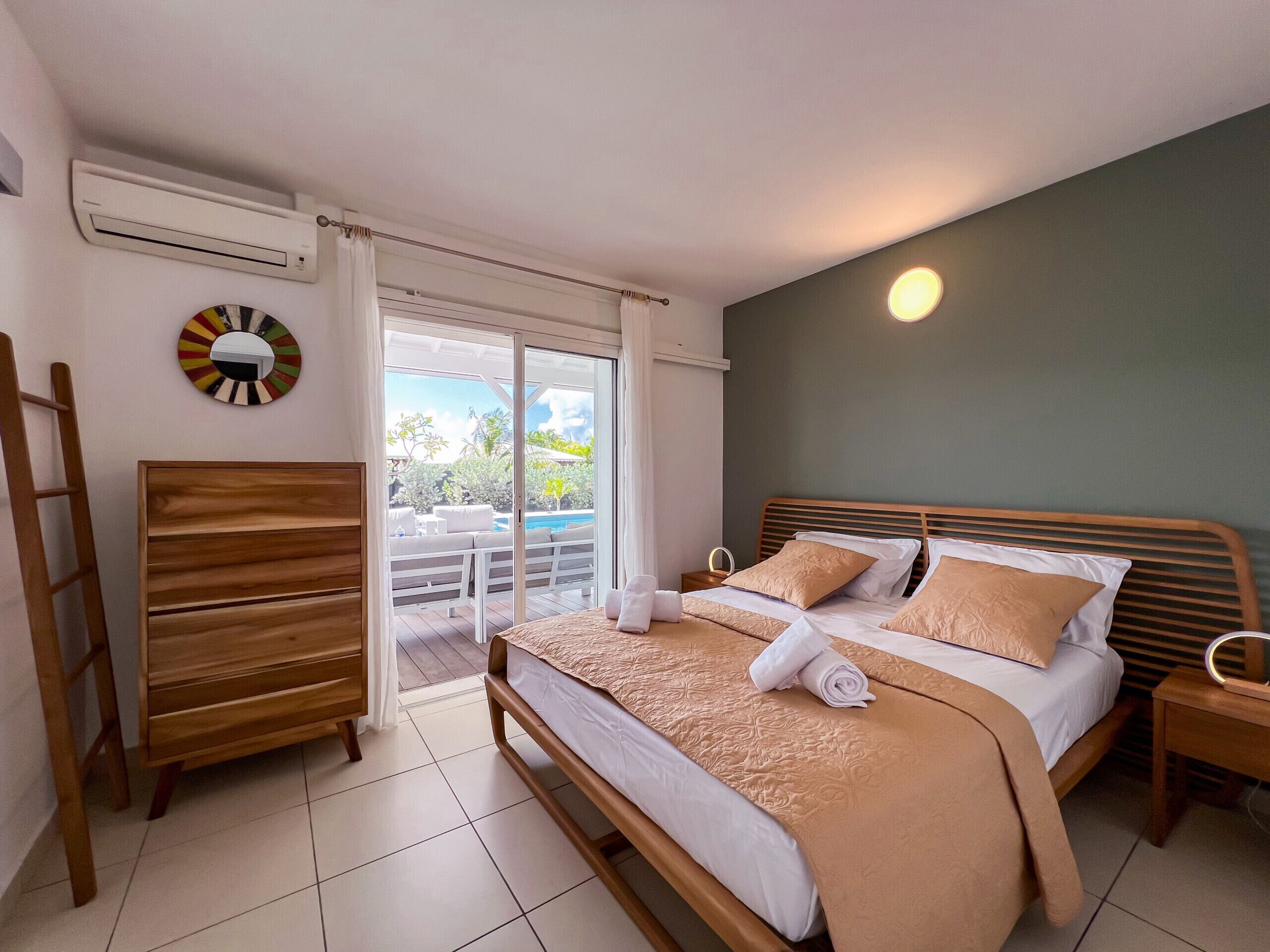 A bedroom with a bed, wooden headboard, a bedside table with a lamp, a mirror, and a view of an outdoor pool area.