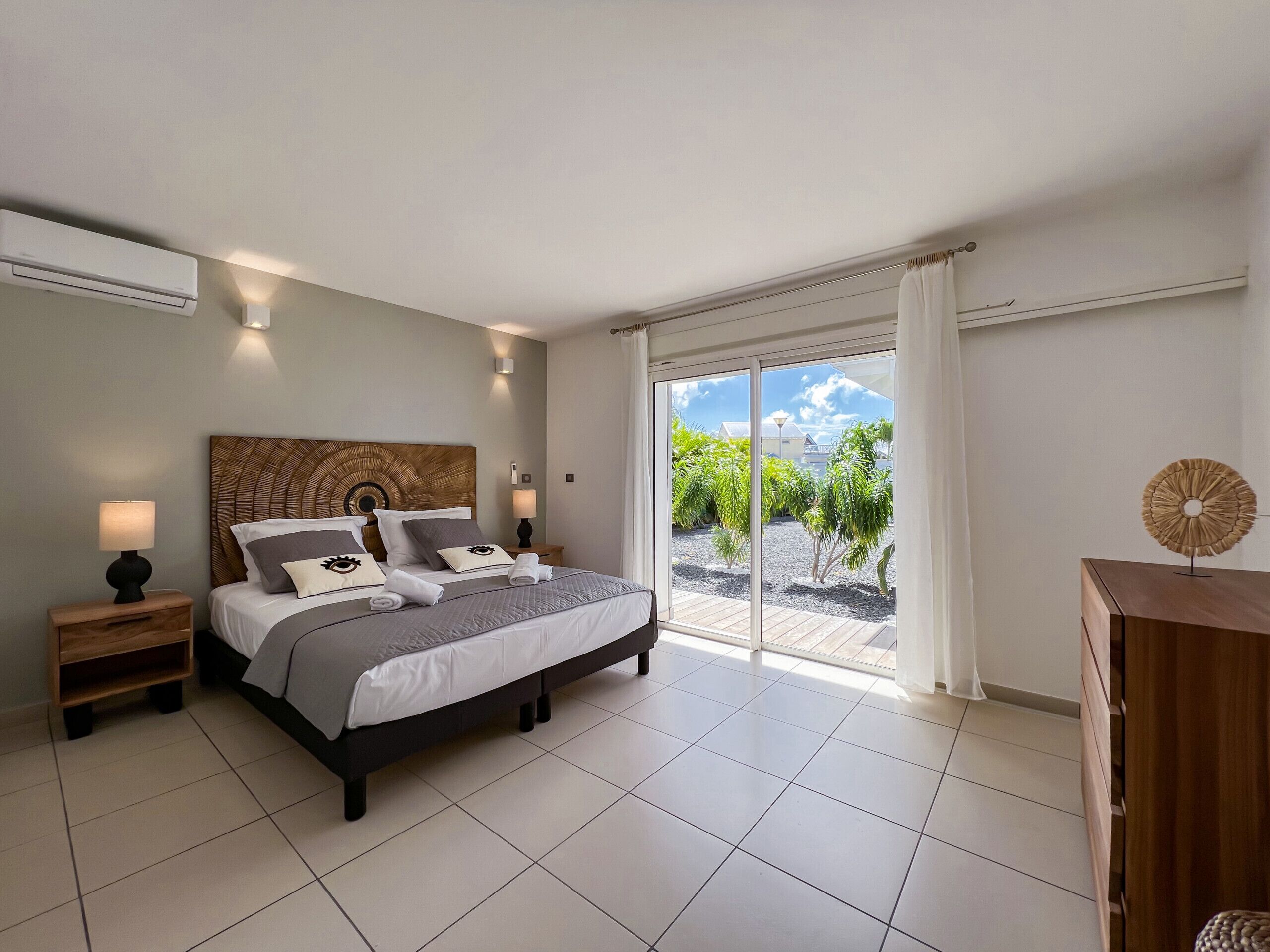 A modern bedroom with a large bed, a nightstand, a dresser, and a view of a garden through a sliding glass door.