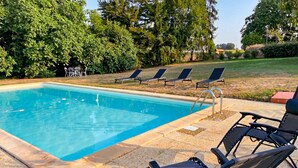 Een rechthoekig zwembad met helderblauw water, omringd door een stenen terras. Vier ligbedden staan langs het zwembad uitgelijnd, en er is een kleine tafel met stoelen op de achtergrond.