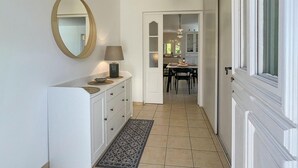 Een gang met een witte dressoir, een spiegel, een lamp en een kamerbreed tapijt dat leidt naar een eetgedeelte met een tafel en stoelen.