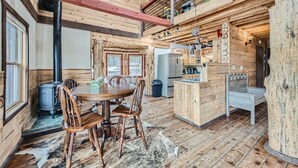 Interior rústico de cabana com mesa de jantar de madeira, cadeiras, área de cozinha e um mezanino.