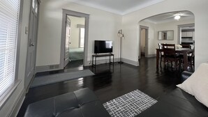 Uma sala de estar espaçosa com televisão de tela plana, uma mesa de jantar com cadeiras e uma cozinha ao fundo.
