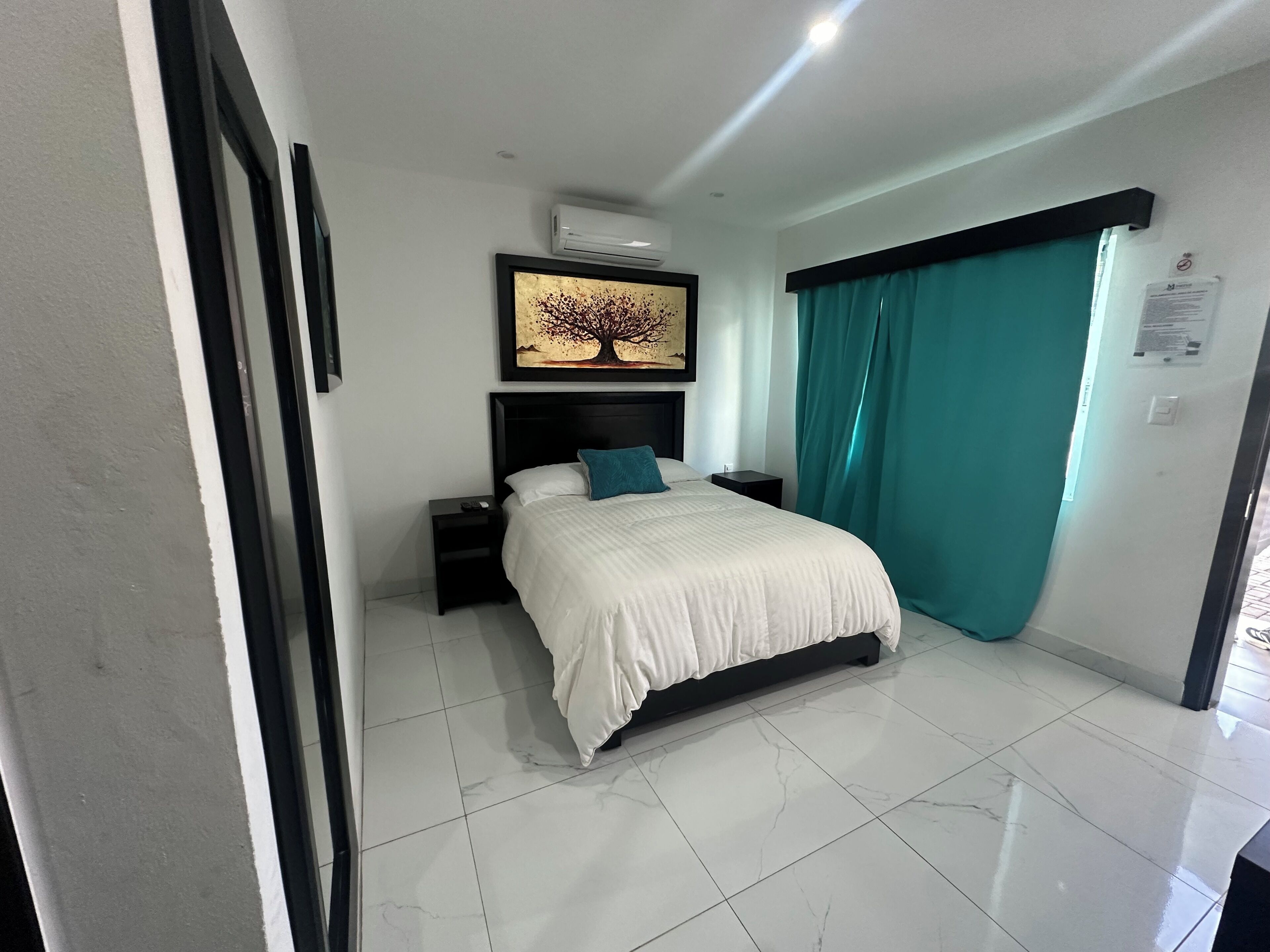A bedroom with a bed, a nightstand, a wall-mounted air conditioner, and a window with teal curtains.