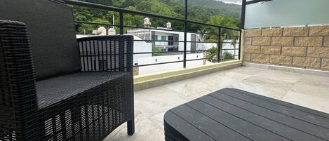 Un balcón con mobiliario de mimbre, vista a un edificio blanco y una ladera verde.