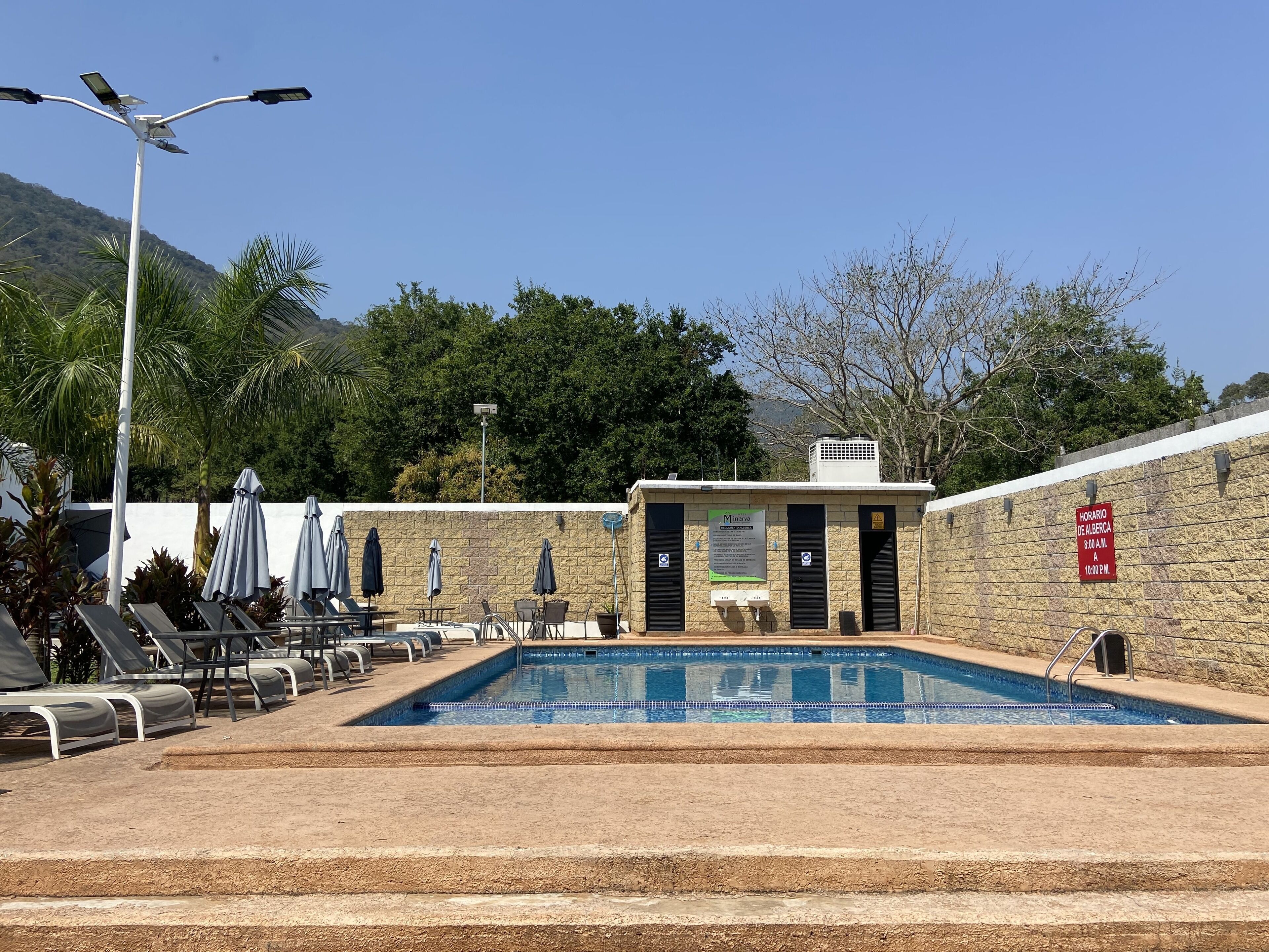 A swimming pool with lounge chairs and umbrellas, a stone wall, and a sign indicating the pool's operating hours.