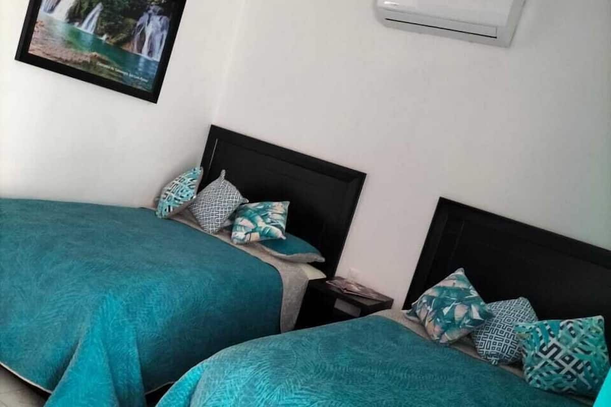 Ein Zimmer mit zwei Betten, jeweils mit türkisfarbenem Bettzeug und gemusterten Kissen, einem gerahmten Bild an der Wand und einer Klimaanlage.
