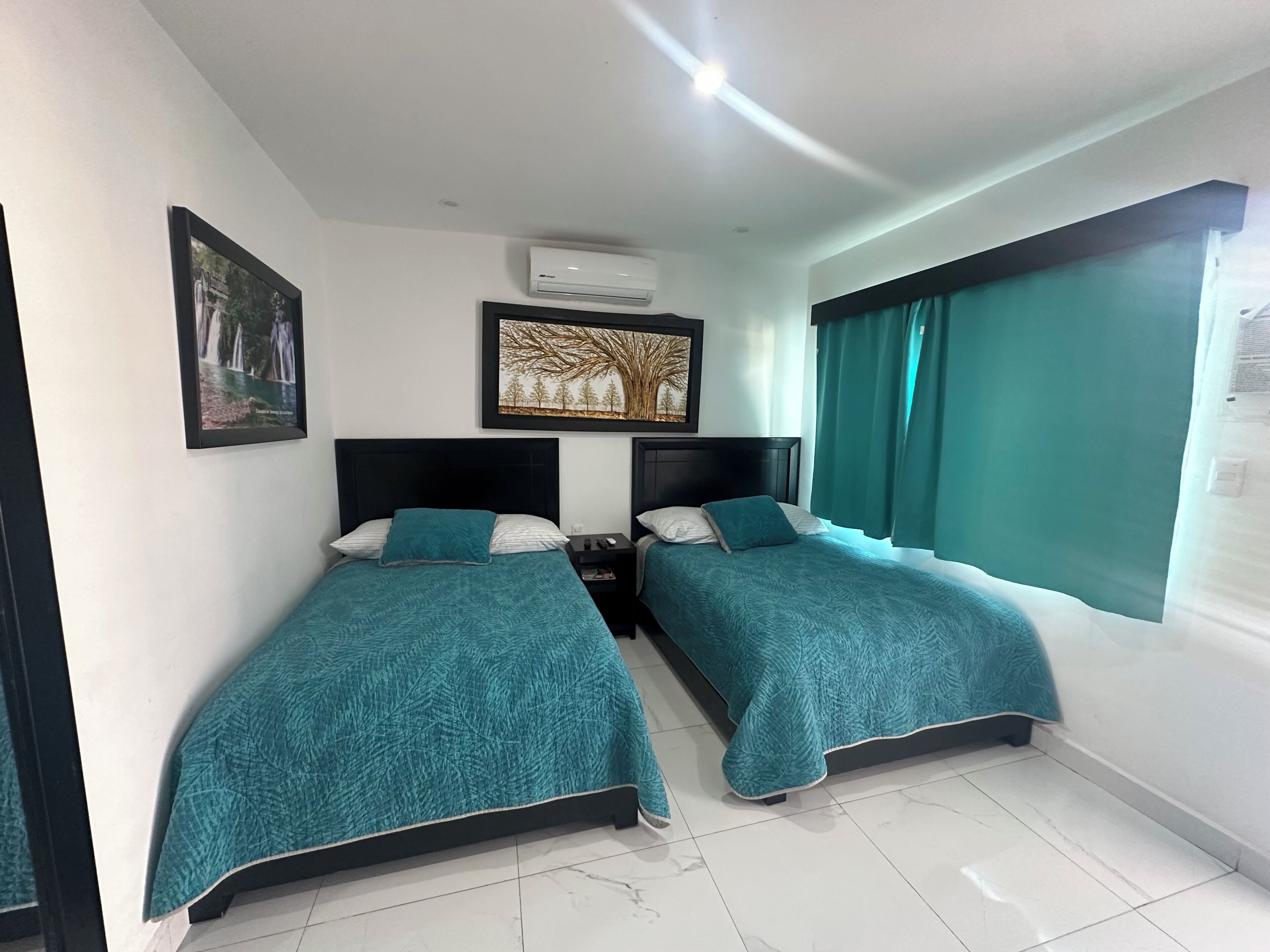 A hotel room with two single beds, a nightstand, a wall-mounted air conditioner, and a window with curtains.