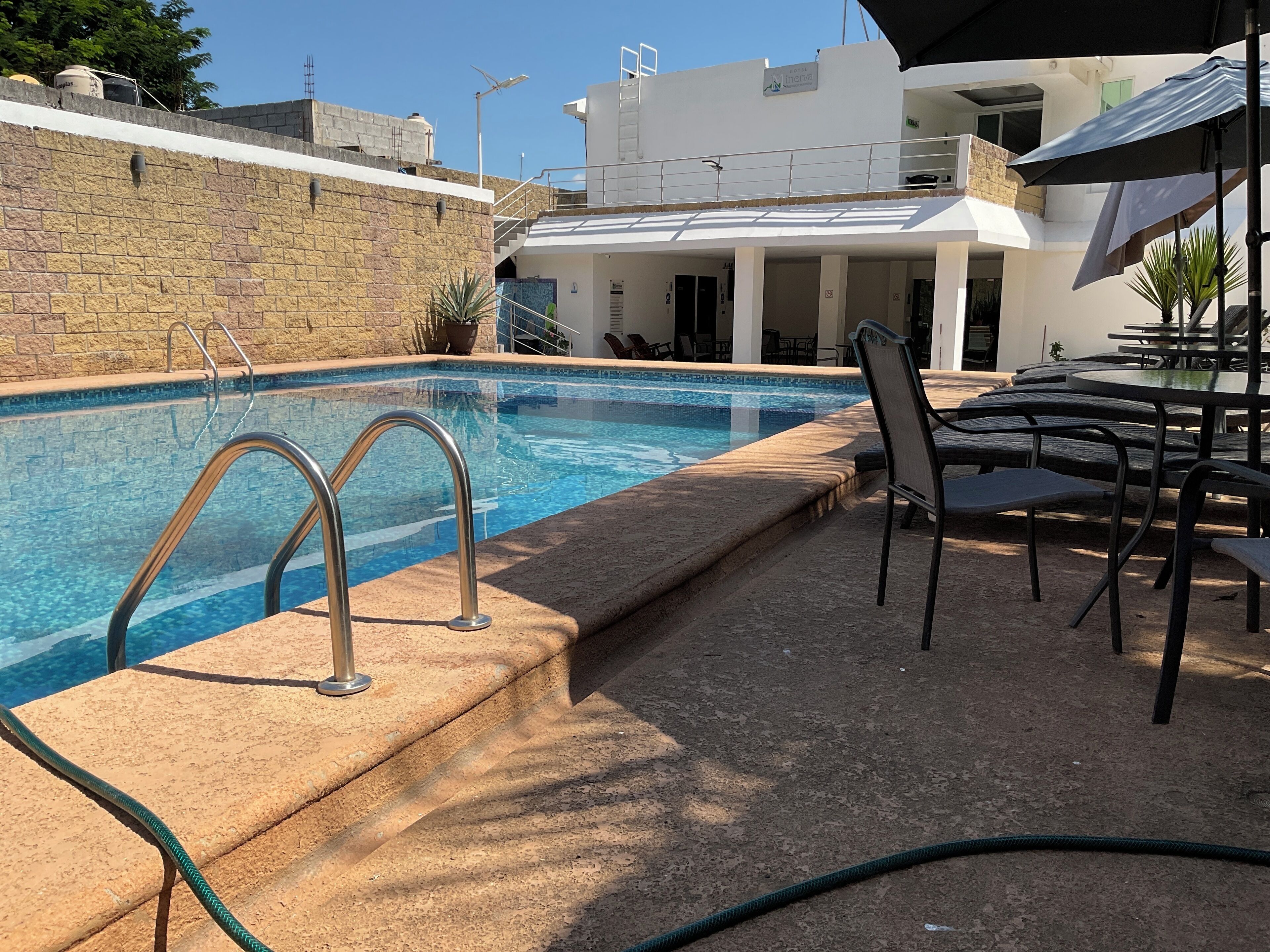 Una piscina con escalones metálicos, un patio con mesas y sillas, y un edificio con balcón al fondo.