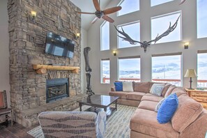 A spacious living room with a stone fireplace, large windows, a ceiling fan, and a mounted deer skull. - Dinosaur Point Lodge (Garden City)