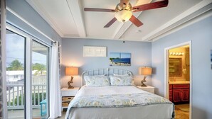 Un dormitorio con cama, dos mesitas de noche, ventilador de techo, un cuadro y vistas al exterior.