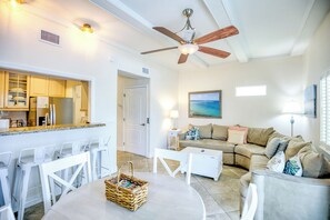 A modern living room with a sofa, coffee table, and chairs. There is a kitchen area with a refrigerator and cabinets. A ceiling fan and a painting are also visible. - Florida Keys, private wellness enjoyment with private hot tub and plunge pool (Marathon)