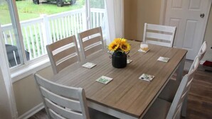Una mesa de comedor con cuatro sillas blancas, un jarrón con girasoles y una ventana que ofrece vista a un césped y un vehículo.