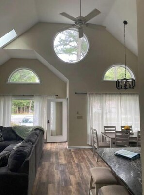 A spacious room with a high ceiling, a ceiling fan, and two large windows providing natural light. There is a dining area with a table and chairs, and a sofa in the living area.