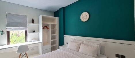 A bedroom with a large bed, a built-in wardrobe, a desk with a chair, and a window with blinds.