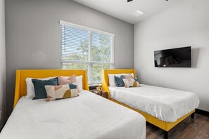 Ein Hotelzimmer mit zwei Betten, einem großen Fenster mit Jalousien, einem an der Wand montierten Flachbildfernseher und einem kleinen Tisch mit einer Lampe.