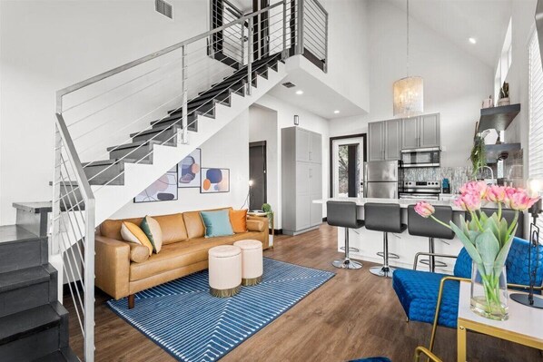 Eine moderne Wohnung mit Treppe, einem Wohnbereich mit Sofa und Hocker, einer Küche mit Barhocker und einem Essbereich mit einem Blumenstrauß.