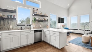 Una cocina moderna con armarios blancos, lavavajillas y una isla grande. Hay ventanas con persianas, una escalera y un televisor de pantalla plana montado en la pared.