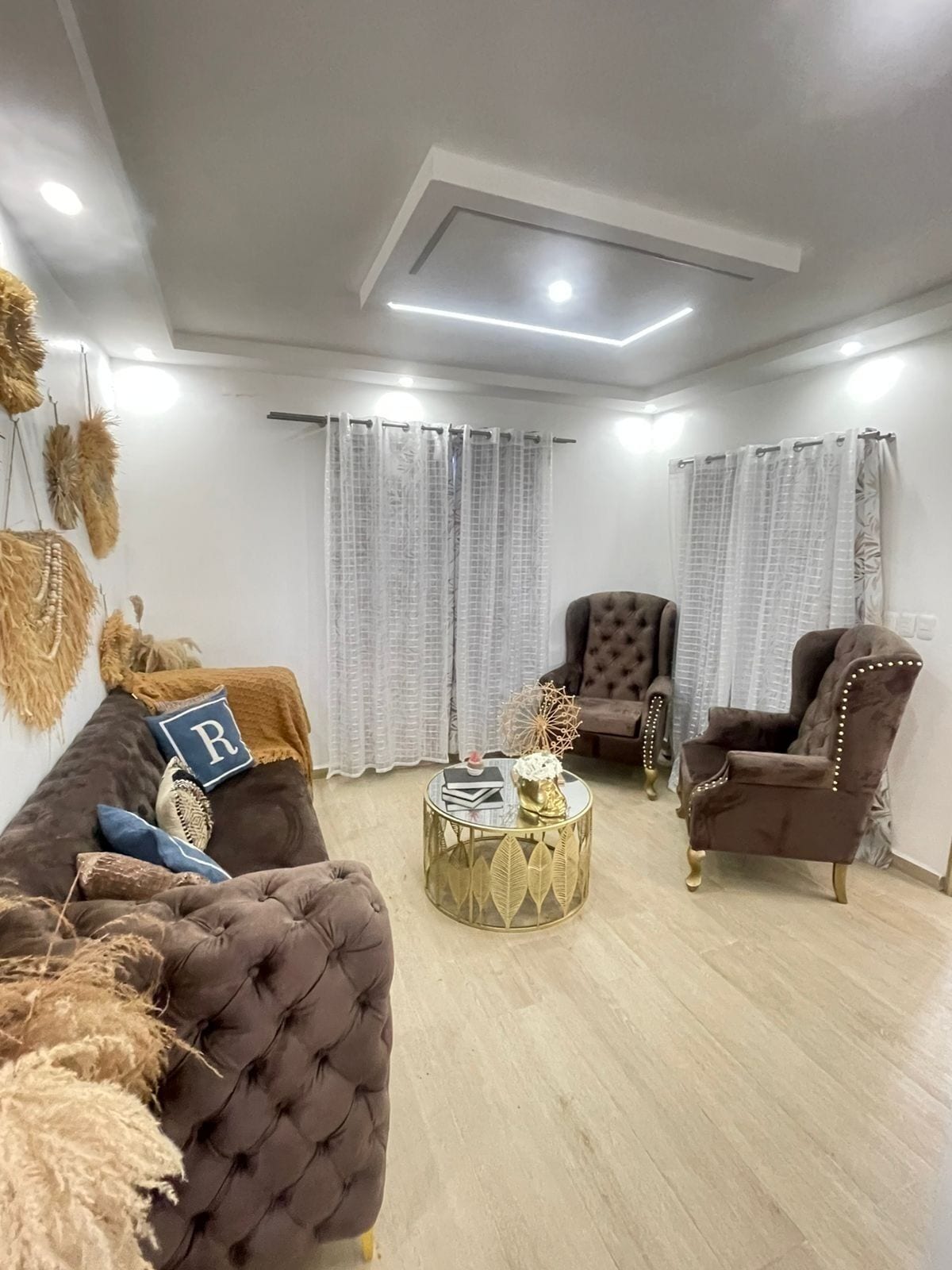 Un salón moderno con un sofá capitonado, dos sillones, una mesa de centro redonda y elementos decorativos como un cojín con la letra 'R' y plantas secas.