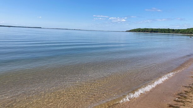 A calm beach with clear water, a sandy shore, and a distant green hill.