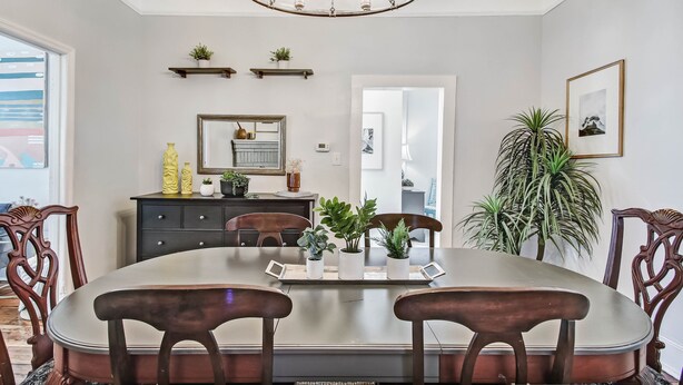 A dining area with a large oval table, wooden chairs, a chandelier, potted plants, a mirror, and a framed picture on the wall.
