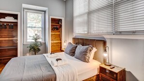 A modern bedroom with a large bed, wooden shelving, a potted plant, and a window with blinds.