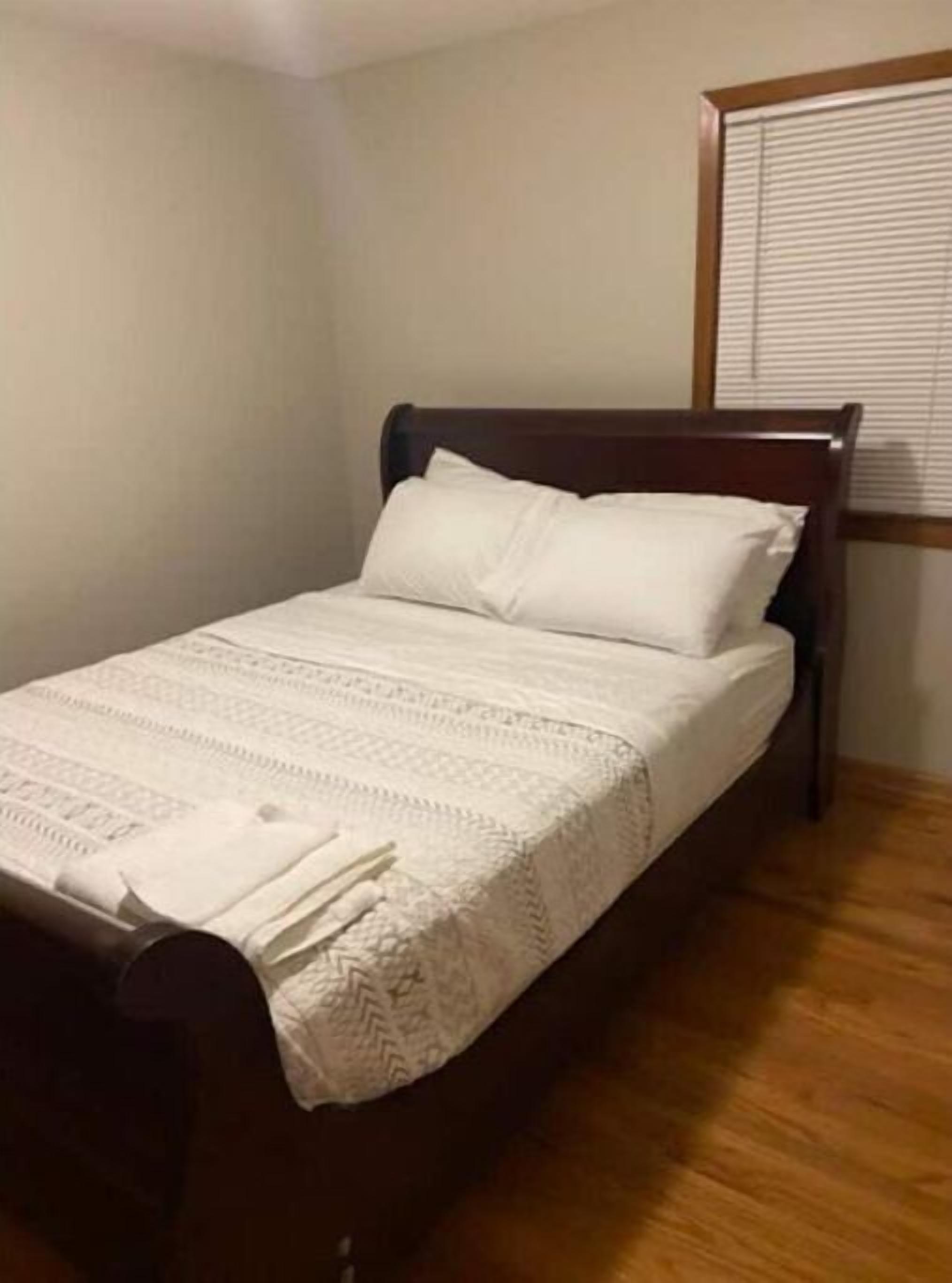 Een eenpersoonsbed met houten frame en witte beddengoed in een kamer met houten vloer en een raam met zonwering.