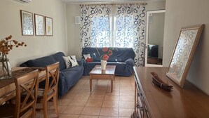 Ein Wohnzimmer mit einer Couch, einem Couchtisch und Sesseln. Im Hintergrund befindet sich ein Fernseher und ein Fenster mit gemusterten Vorhängen.