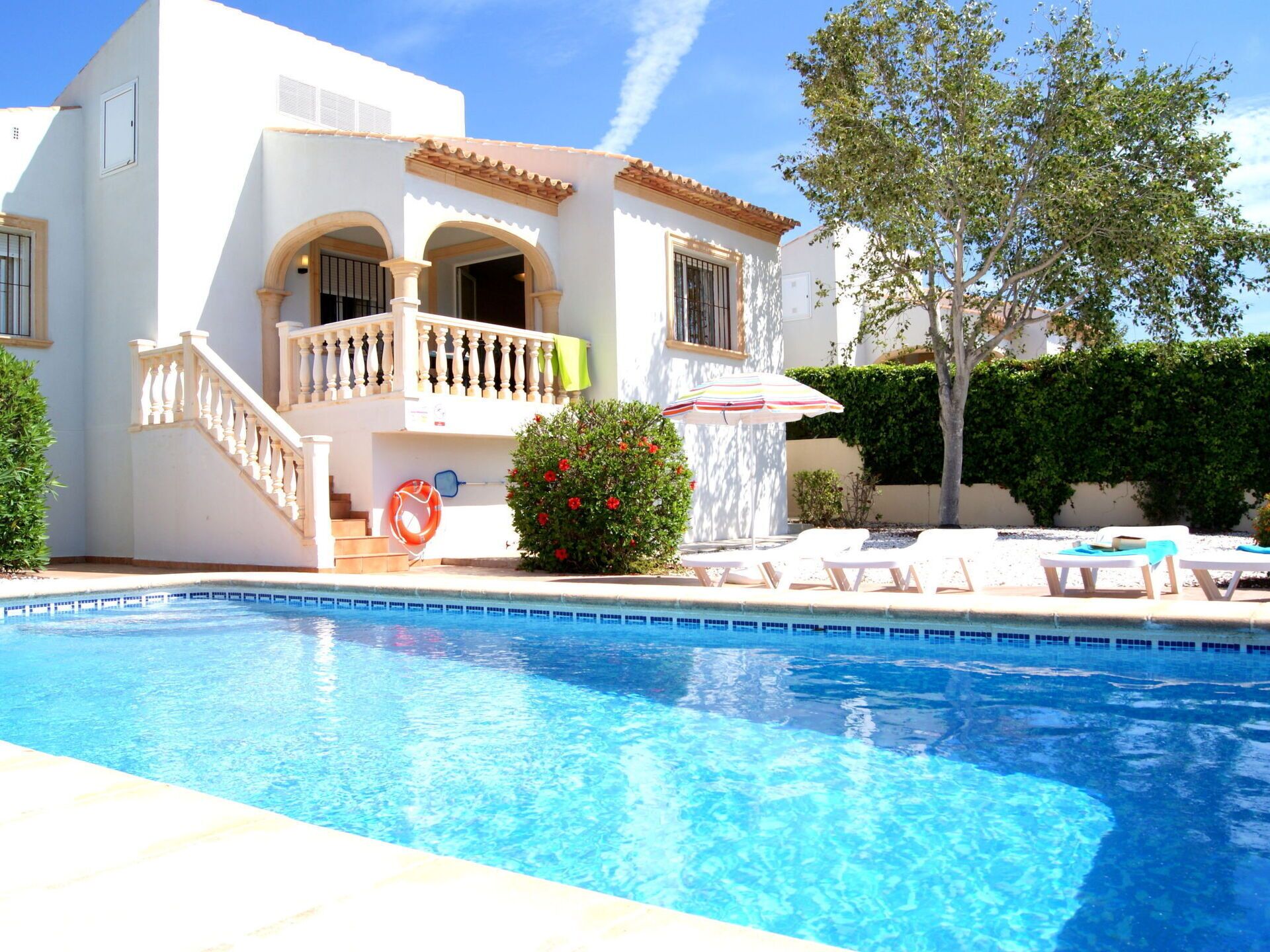A poolside view of a white building with a balcony, a lifebuoy, and lounge chairs.