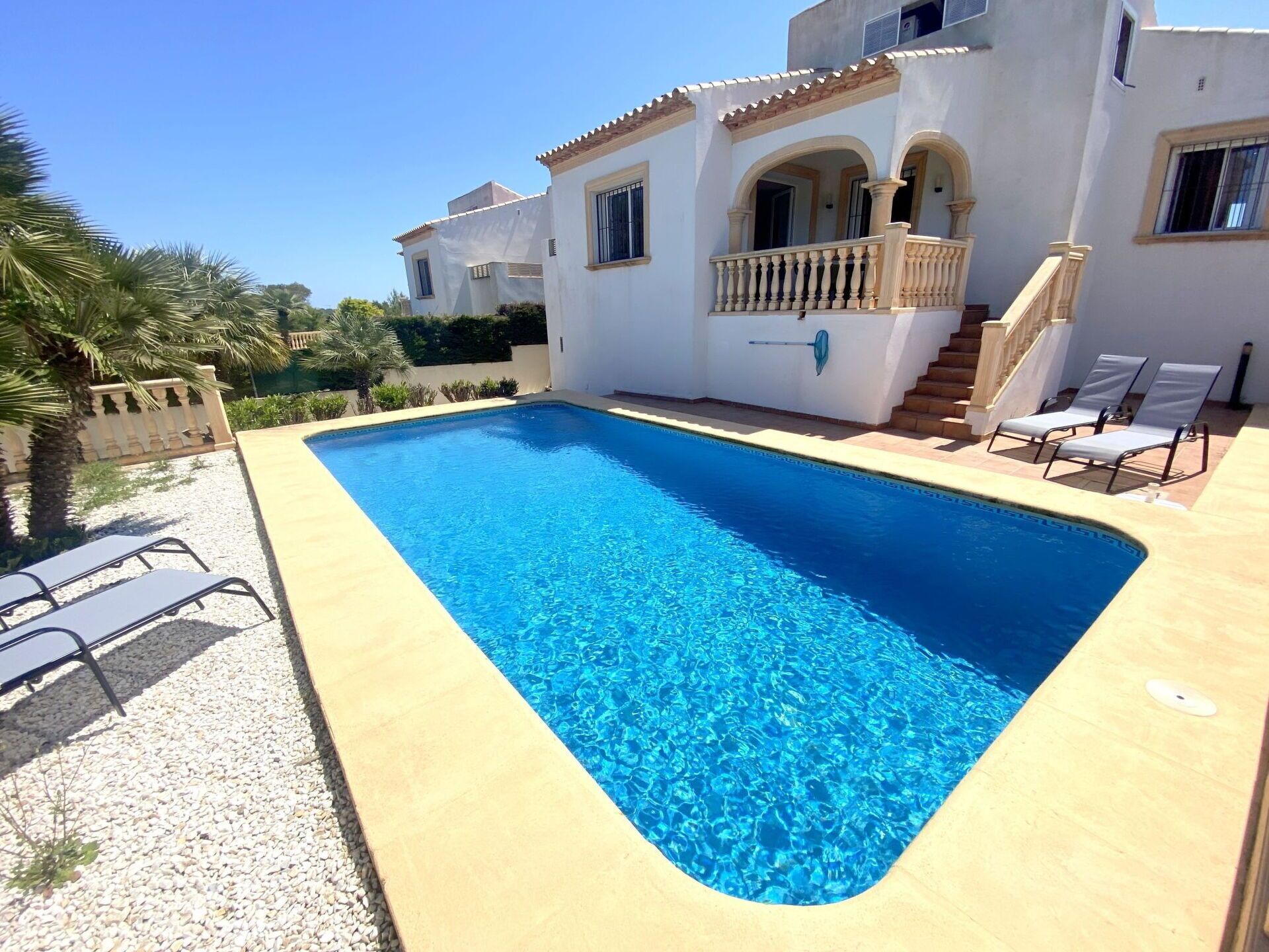 A rectangular swimming pool with clear blue water, surrounded by a light-colored deck. There are two lounge chairs on one side and a staircase leading to a balcony with a railing.