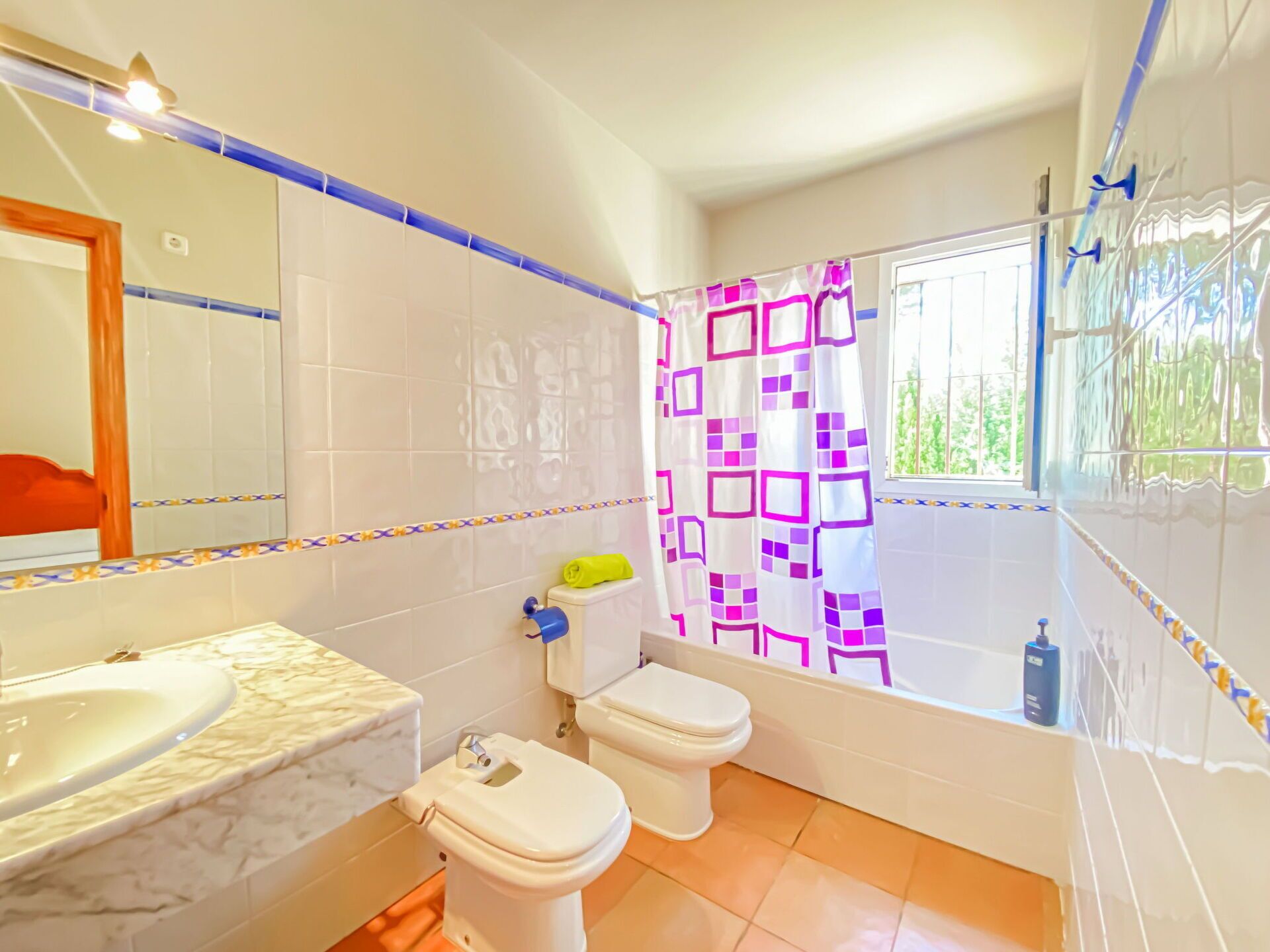 A bathroom with a white sink, two toilets, a shower curtain with a geometric pattern, and a window with a view of greenery.