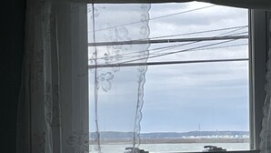 A room with a large window overlooking a waterfront area, featuring a wooden dock and a distant view of a city skyline. - Oysterhaven Cottage with partial Water views! New balcony! Close to Fairgrounds! (Chincoteague)