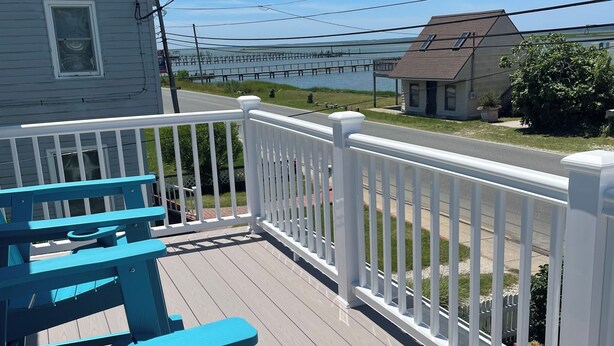 A wooden deck with white railings, a blue bench, and a view of a dock and houses across the water. - Oysterhaven Cottage with partial Water views! New balcony! Close to Fairgrounds! (Chincoteague)