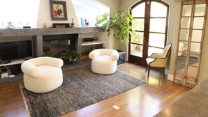 Un salon avec une cheminée, deux fauteuils blancs, une grande plante et une vue à travers une porte vitrée.