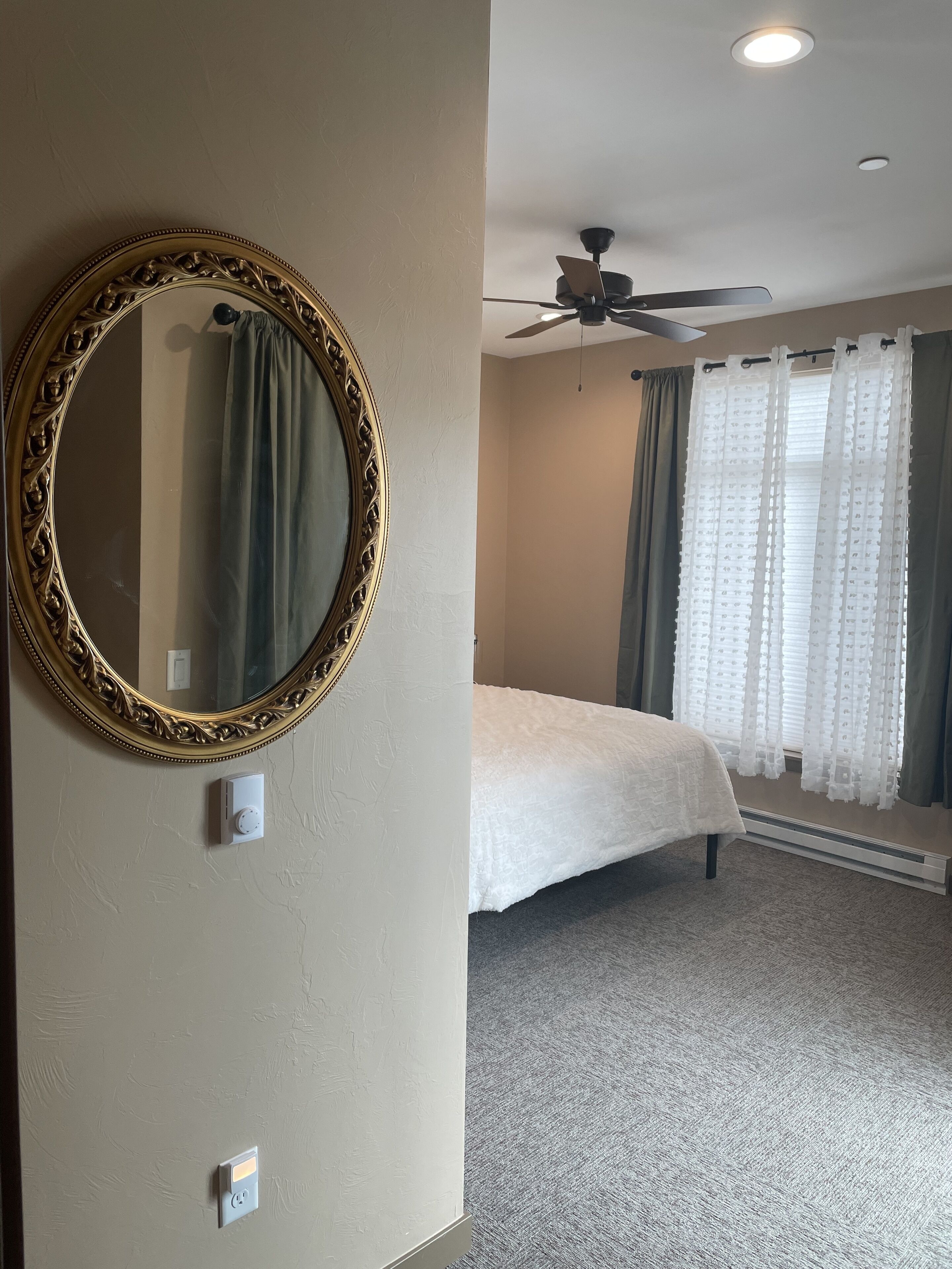 A bedroom with a large ornate mirror, a bed with a white comforter, a ceiling fan, and sheer curtains.