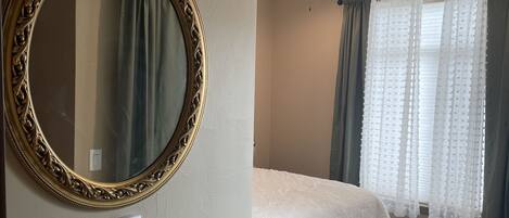 A bedroom with a large ornate mirror, a bed with a white comforter, a ceiling fan, and sheer curtains.