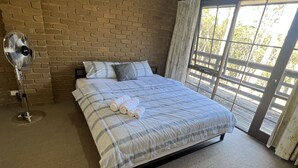 A bedroom with a brick wall, a bed with a plaid bedspread, a standing fan, and a large window with curtains. - 1 bedroom studio very quiet with farm view in Heathcote junction. (Heathcote Junction)