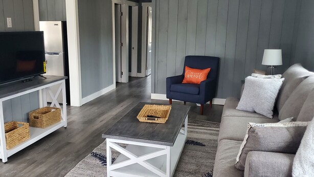 Un salon avec un téléviseur posé sur une console blanche, un fauteuil bleu avec un coussin orange, un canapé gris et une petite table sur laquelle se trouve un panier.