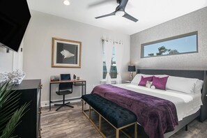 A modern hotel room with a bed, desk, and chair, a ceiling fan, and a window with a city view.