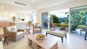 A spacious living area with wicker furniture, a wooden coffee table, and a view of a lush garden through large sliding glass doors. - La Bella Waters 3 (Whitsundays)