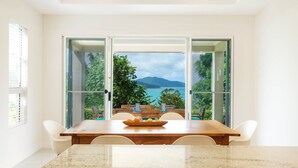 A dining area with a large table and chairs, a bowl of fruit, and a view of a lake and mountains through glass doors. - La Bella Waters 3 (Whitsundays)