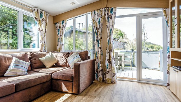 A living room with a brown sofa, large windows, and a view of greenery. - 1 bedroom accommodation in Mullion Cove, Helston (Mullion Cove, Helston)