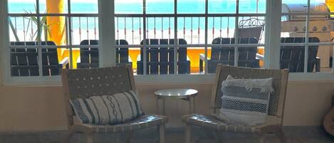 A room with two wicker chairs and a small table, a large window with a view of the beach and ocean, and a grill outside.